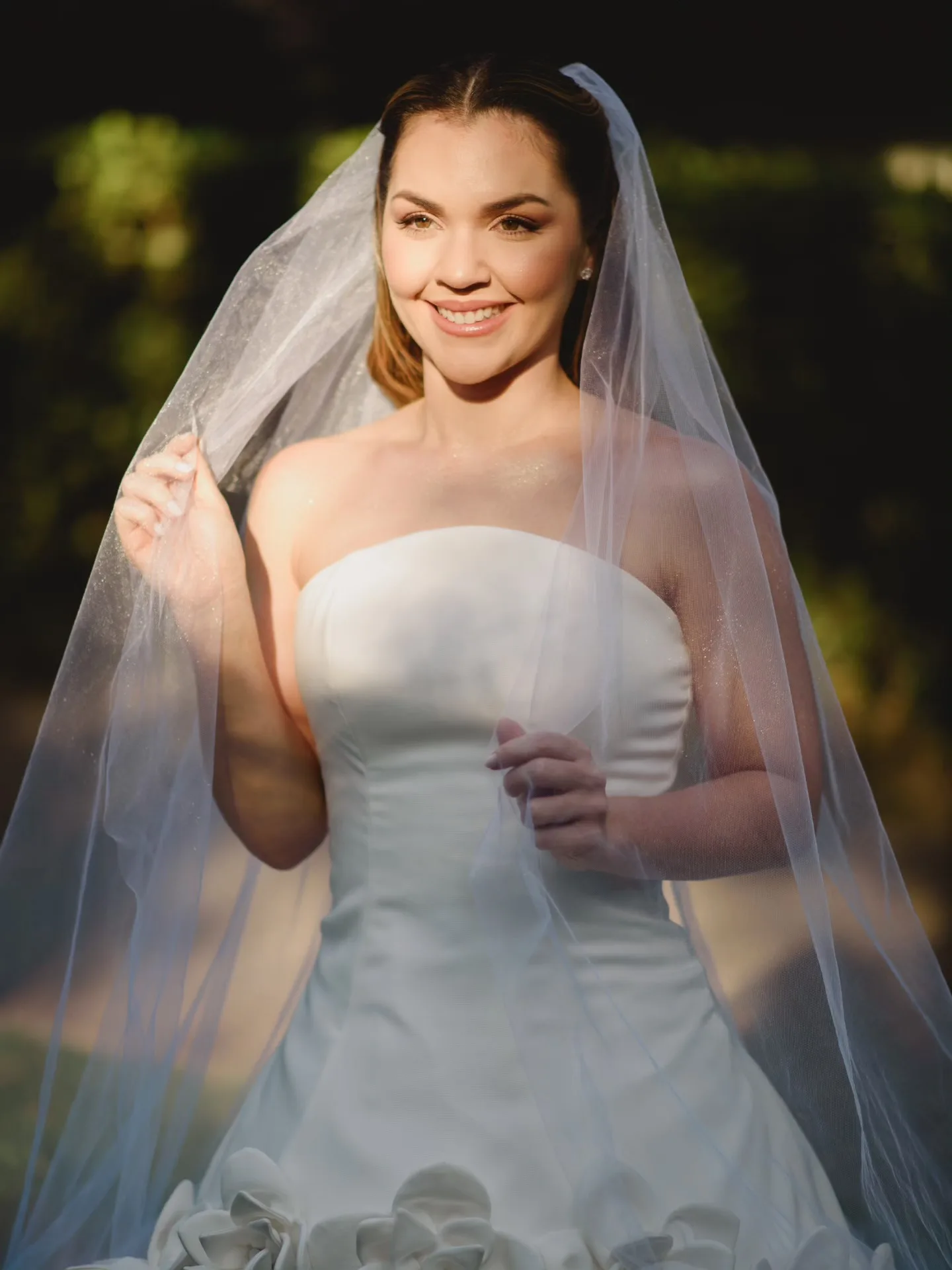 Noiva sorrindo com o véu diante de árvores.