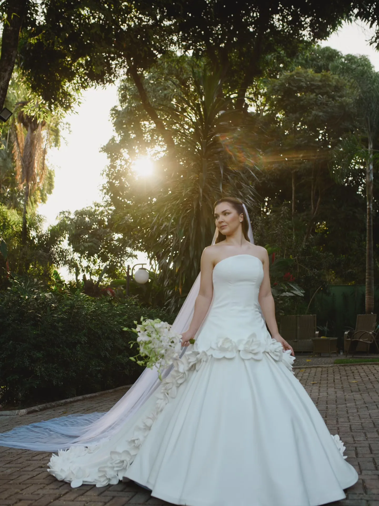 Noiva de vestido longo em um jardim iluminado pelo sol.