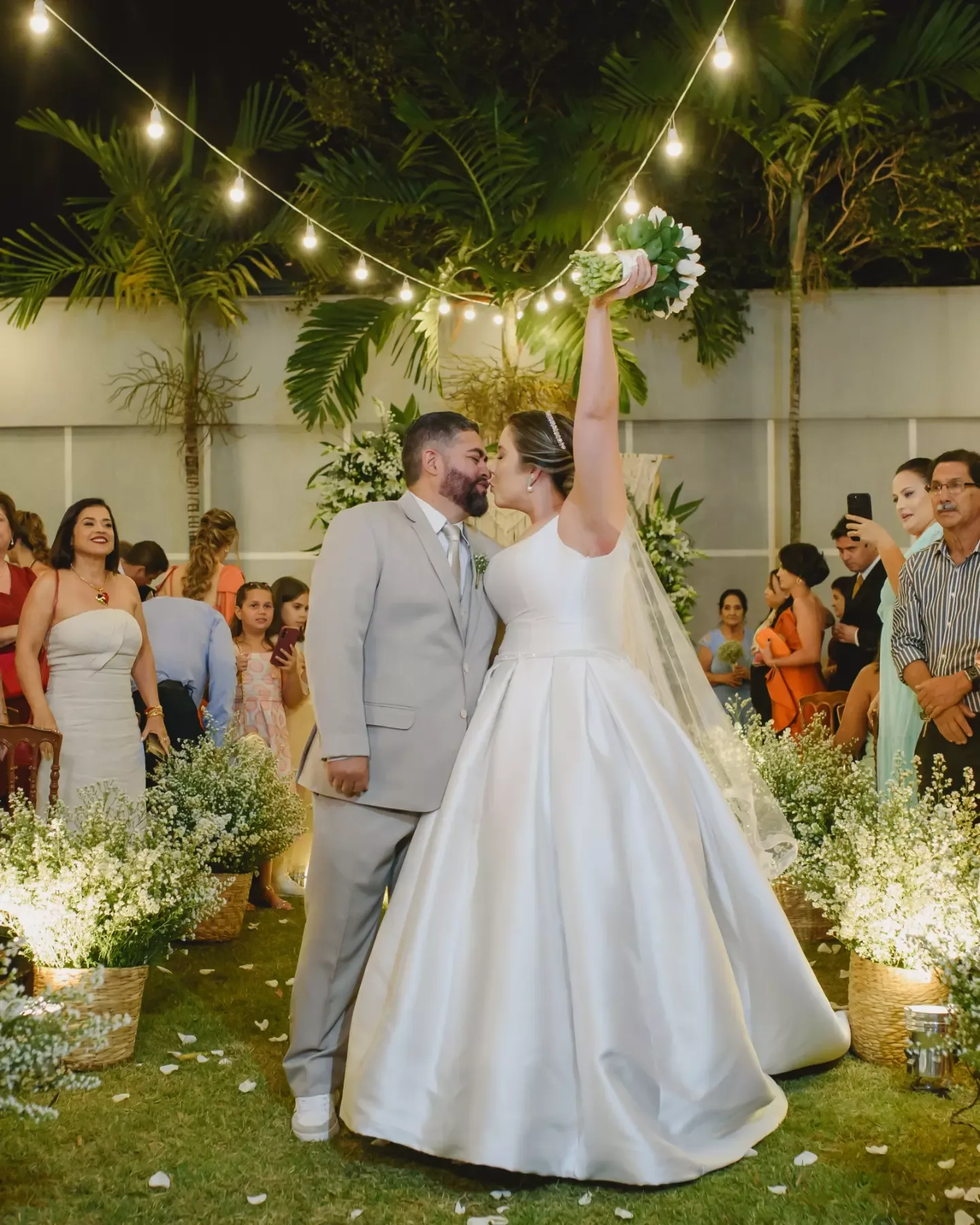 Noivos de mãos erguidas durante uma celebração sob cordões de luz.