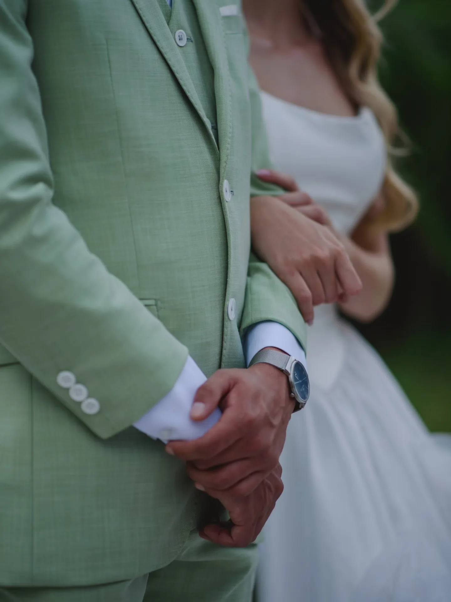 Detalhe das mãos dos noivos, com terno verde e vestido branco.