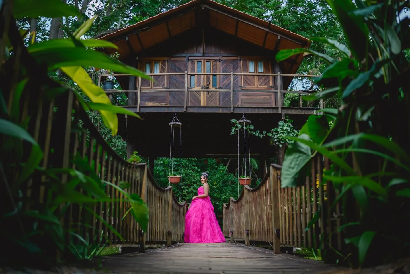 Jovem de vestido rosa diante de uma casa de madeira cercada por árvores.