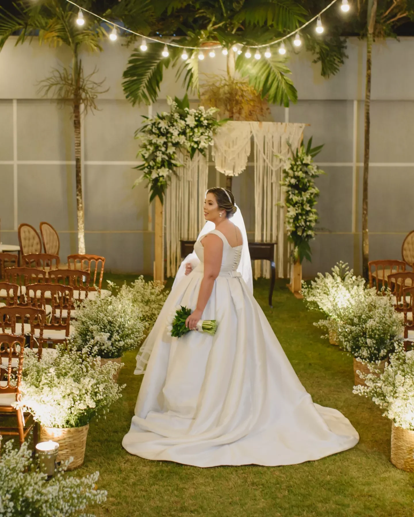 Noiva de vestido longo diante de um arco florido e luzes suspensas.