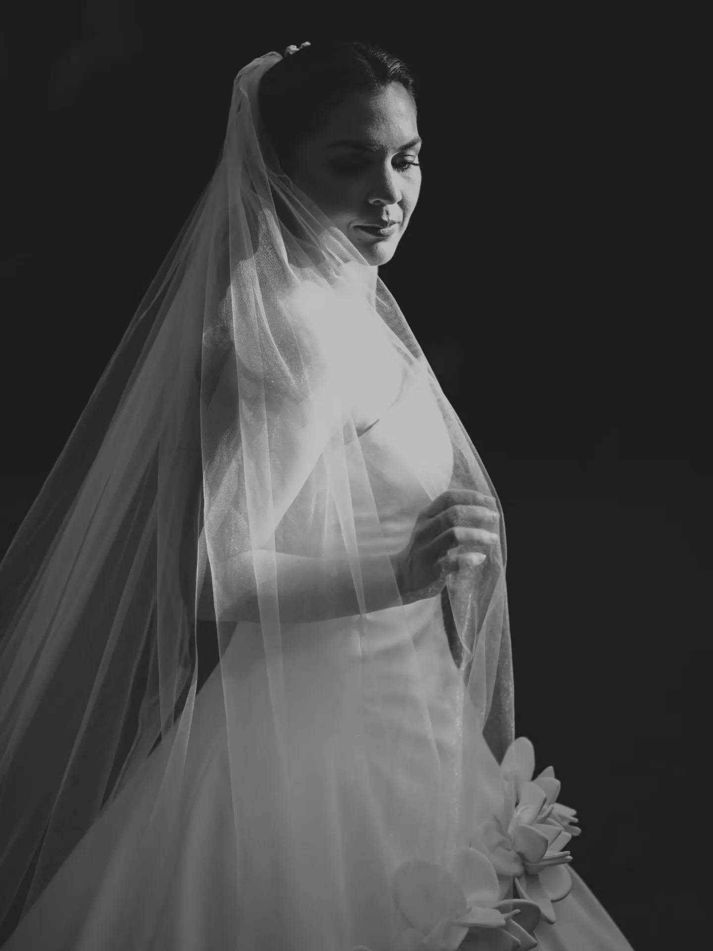 Noiva de perfil junto à janela, com o véu iluminado em preto e branco.