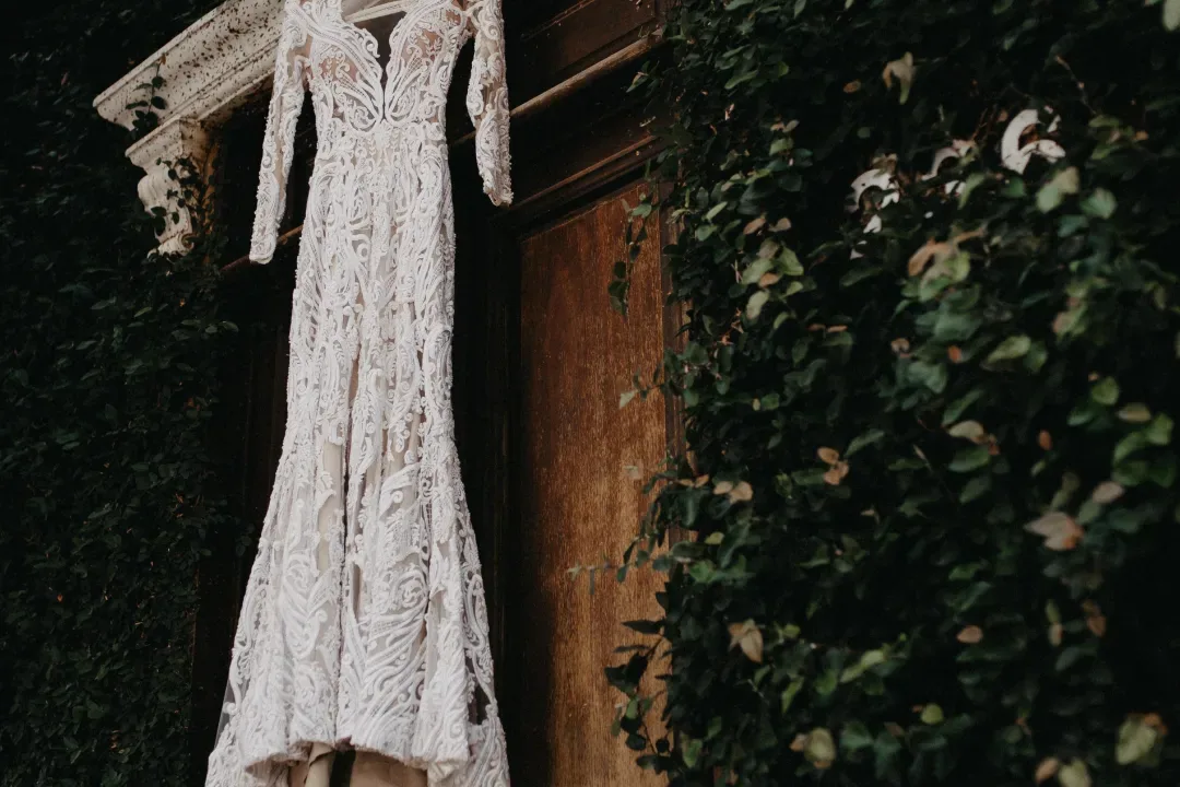 Vestido de noiva pendurado junto a uma porta de madeira e vegetação.