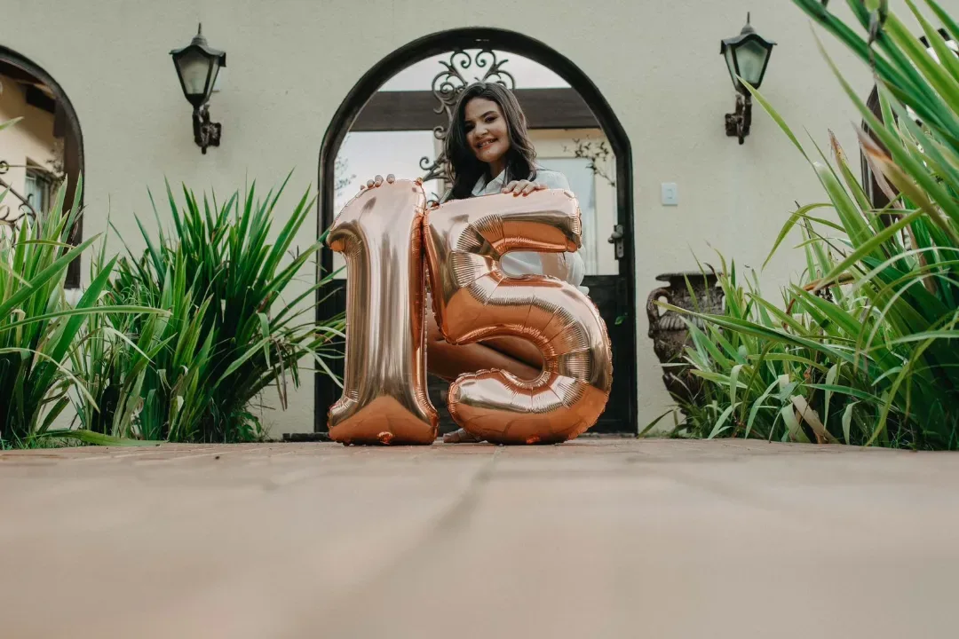 Jovem sentada diante de um arco, segurando balões com o número quinze.
