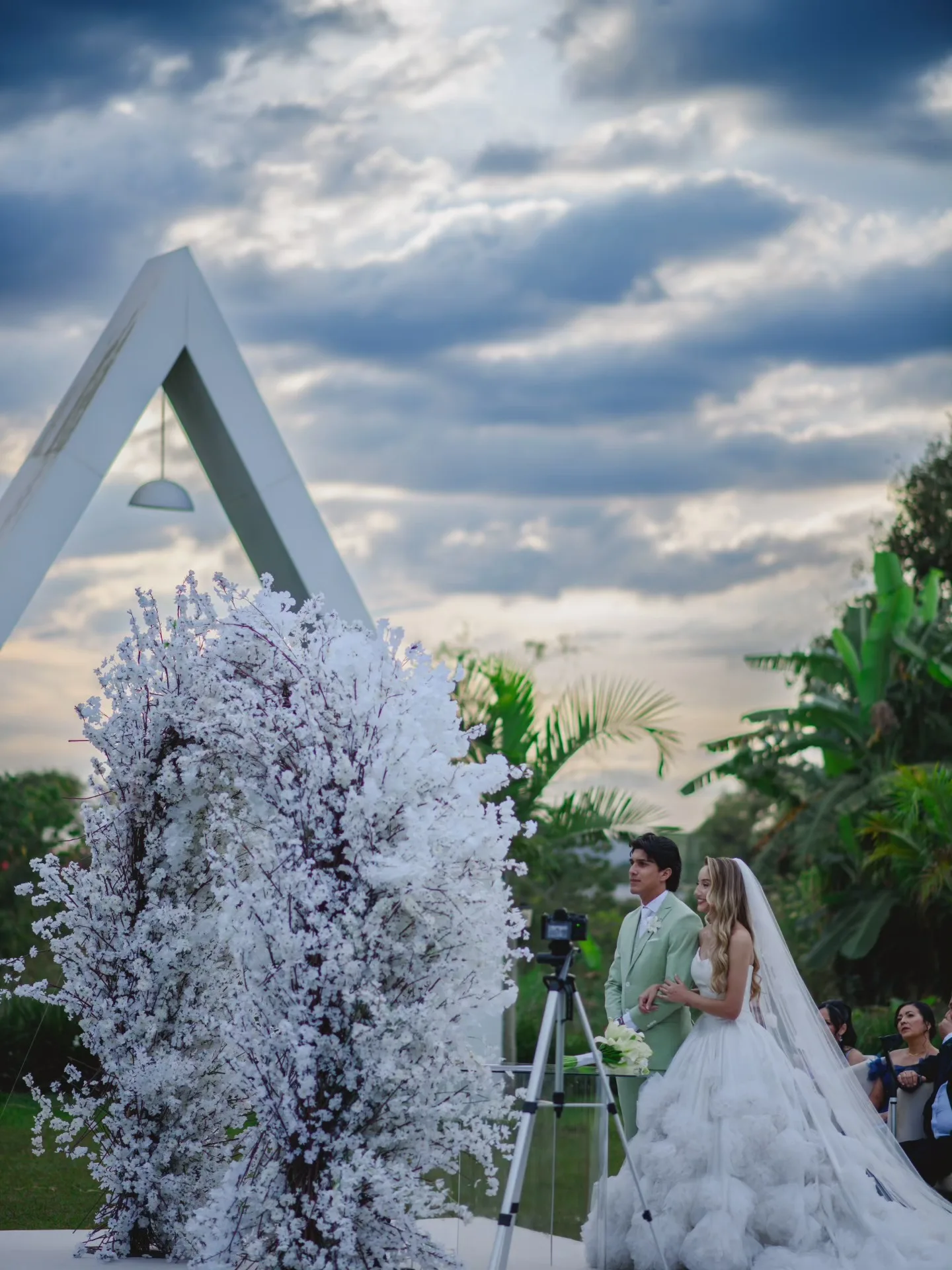 Noivos em um jardim sob um arco branco e céu com nuvens.