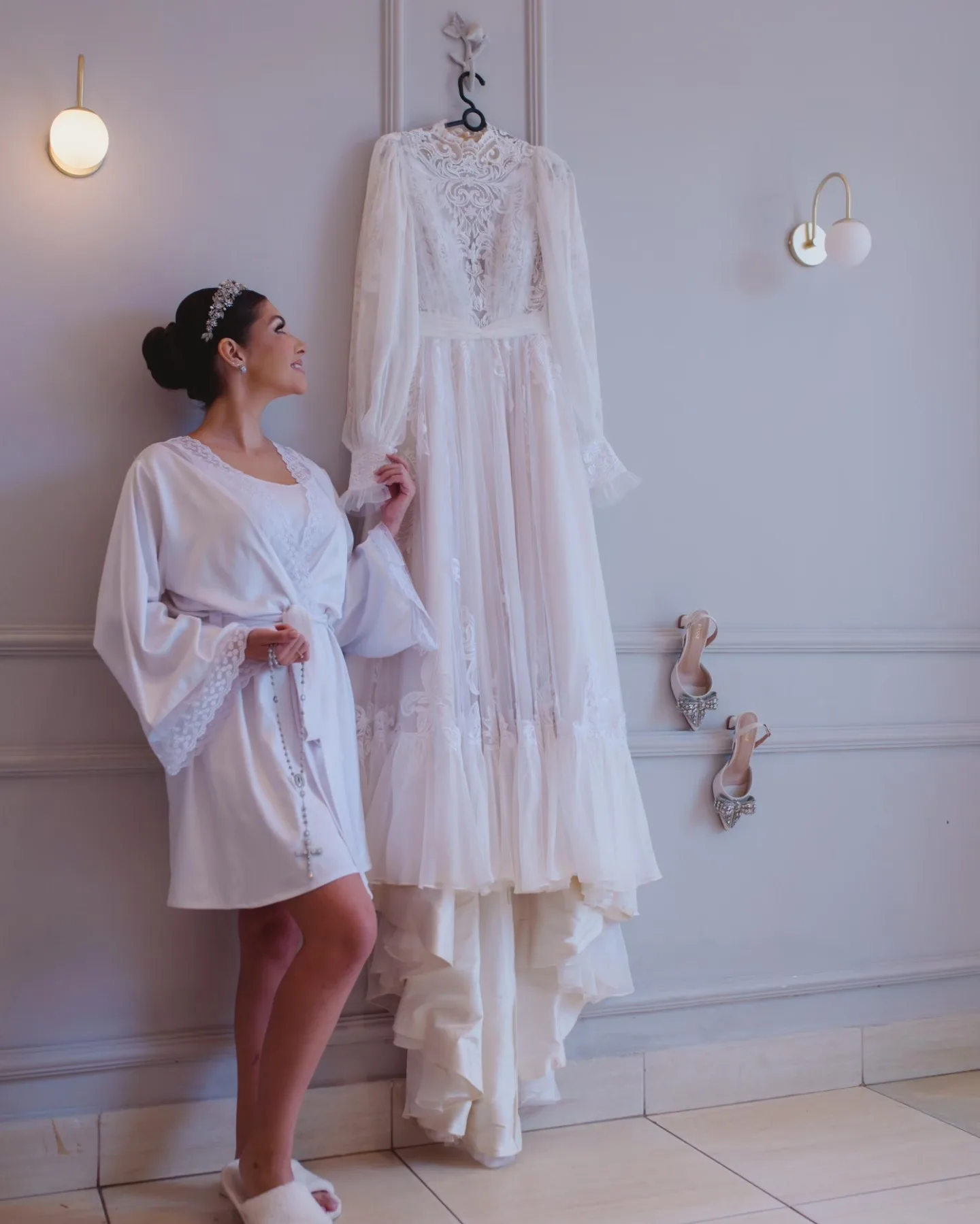 Noiva de robe junto ao vestido pendurado em uma sala clara.