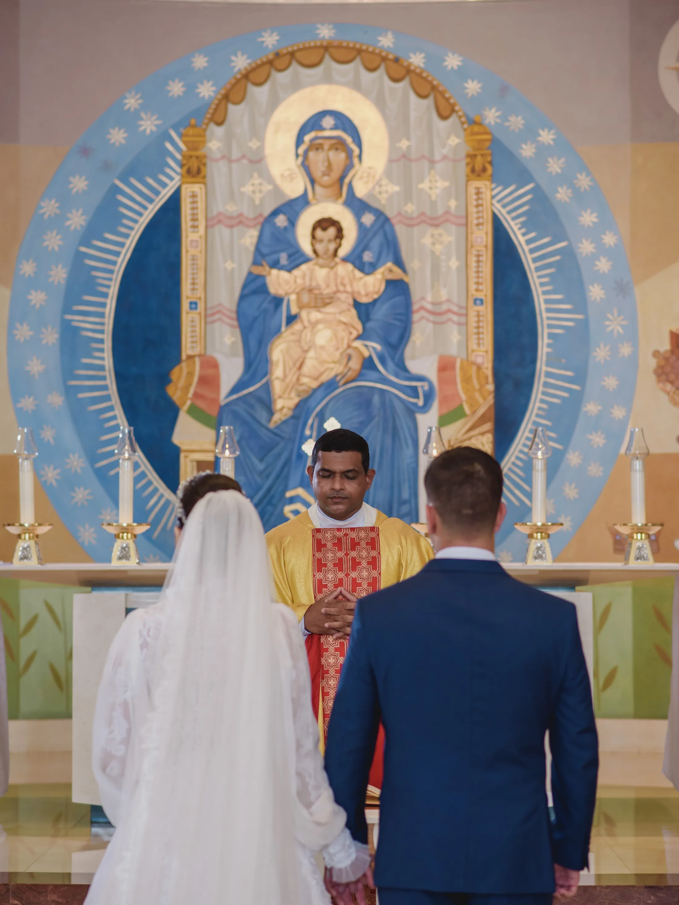 Casal de noivos diante do altar com painel azul e dourado.